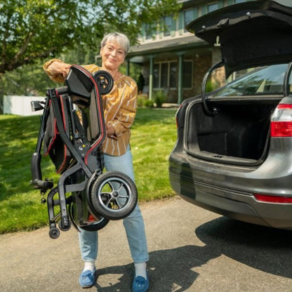 Showing the ScooterPac FeatherFold Mobility Scooter, folded and being lifted by a lady infront of her car boot