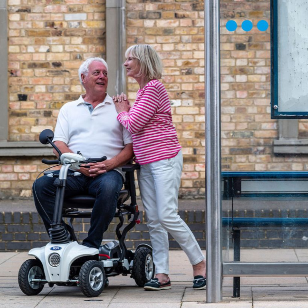 Showing the Maximo Plus with a man sat on the scooter at a bus stop, with his wife leaning on his shoulder smiling.